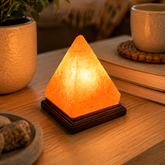 A glowing pyramid salt lamp on a dark wooden base, placed on a wooden table in a cozy, warmly lit living room setting with a plant, books, and a mug in the background.