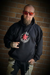 A man stands in front of a brown brick wall wearing a black hoodie with a red and white graphic, black pants with a camouflage pattern on the upper legs and white camouflage on the lower legs, and colorful sneakers.