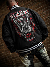 A man viewed from behind wearing a black bomber jacket with a large red, white, and gray graphic on the back, posed against a brick wall.