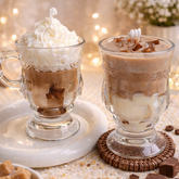 Two glass mugs with hot chocolate, whipped cream, and chocolate pieces on a decorative surface with lights and flowers in the background.