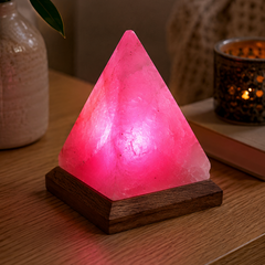 A pink Himalayan salt crystal pyramid lamp glowing on a dark wooden base, placed on a wooden table in a cozy home setting with a plant and candle in the background.