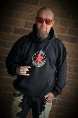 A man stands in front of a brown brick wall wearing a black hoodie with a red and white graphic, black pants with a camouflage pattern on the upper legs and white camouflage on the lower legs, and colorful sneakers.