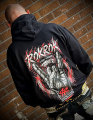 A man viewed from behind wearing a black hoodie with a large red, white, and gray graphic on the back that reads 'ROKROK' and 'STING CHAMPIONSHIP'. He is standing against a brown brick wall.