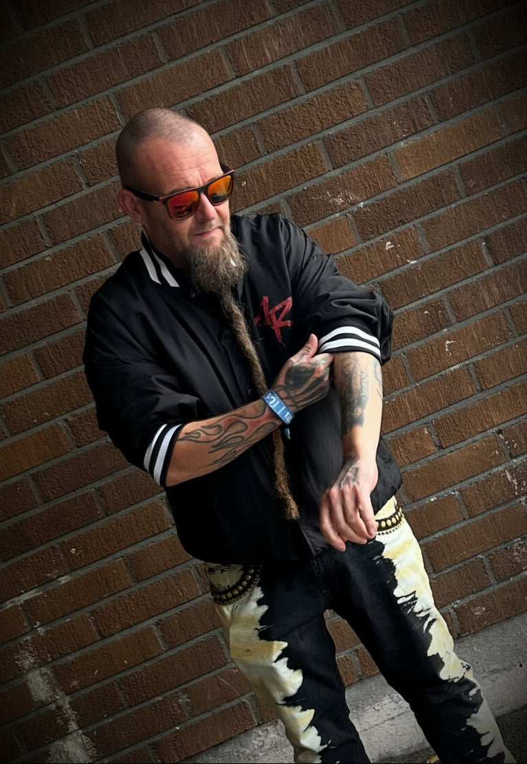 A man with a beard and sunglasses poses against a brick wall, wearing a black bomber jacket, black and beige camouflage pants, and colorful sneakers.