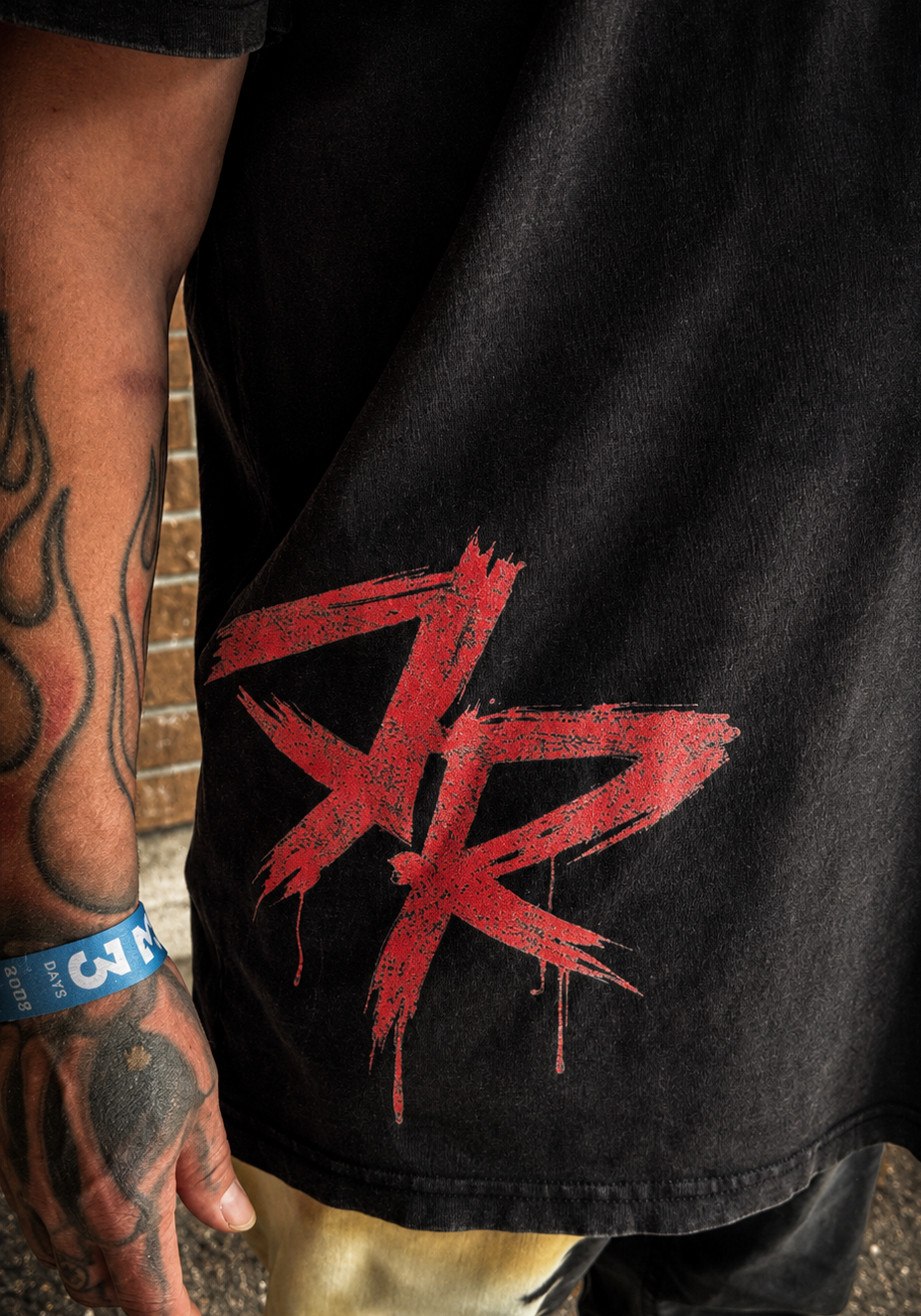 A close-up of a person wearing a black graphic t-shirt with a large red 'DR' logo on the lower front, paired with distressed beige and black pants.
