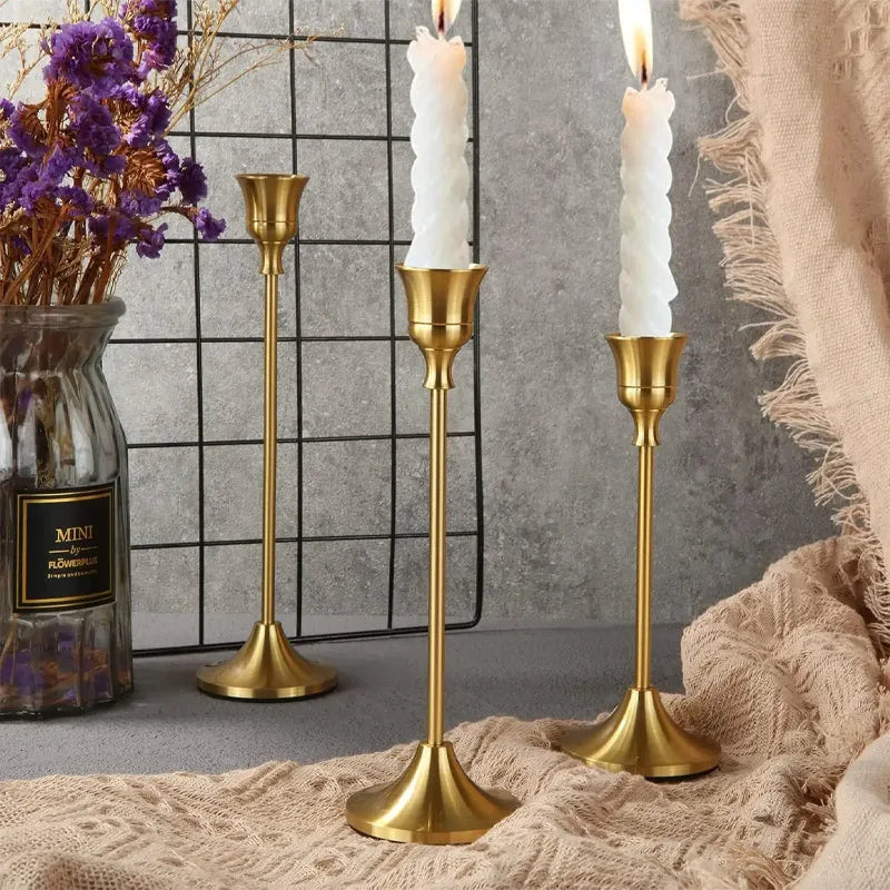 Gold candle holders with white candles on a textured surface with a vase of flowers.