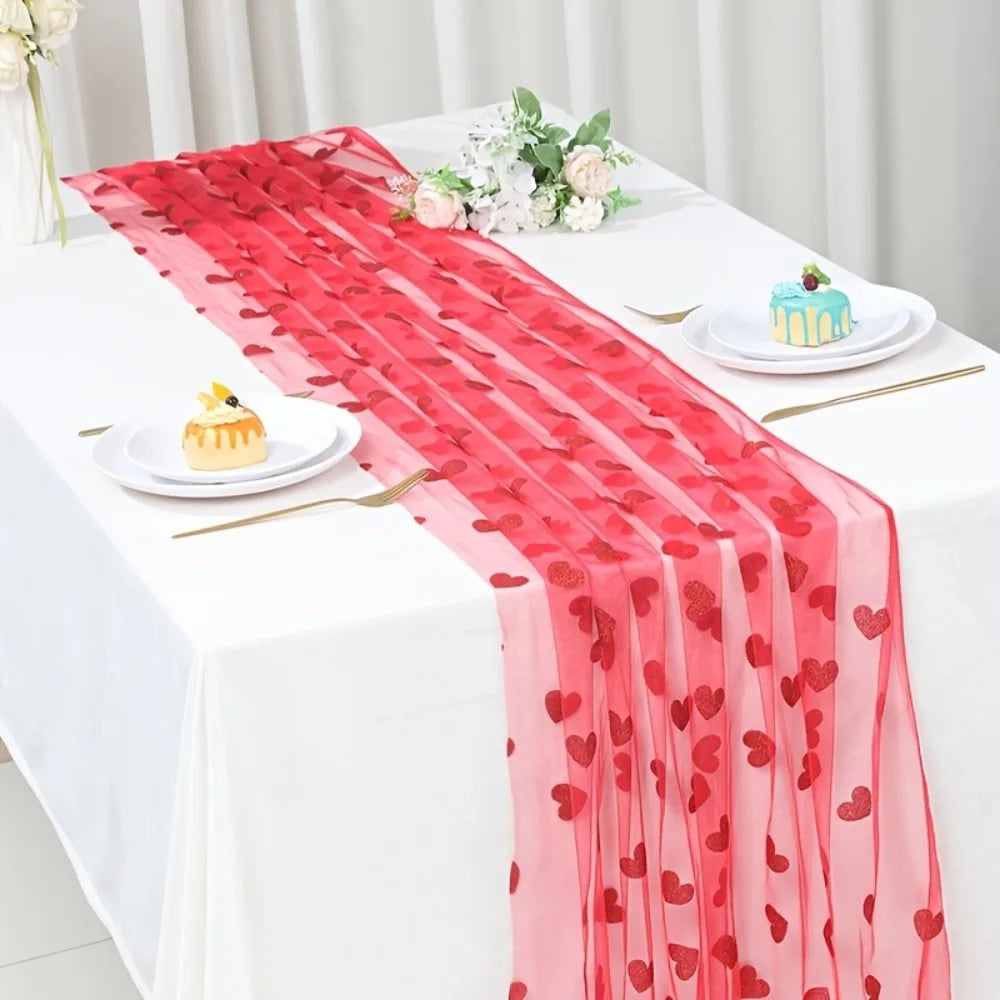 Table setting with a red heart-patterned table runner, plates with desserts, and floral arrangements.