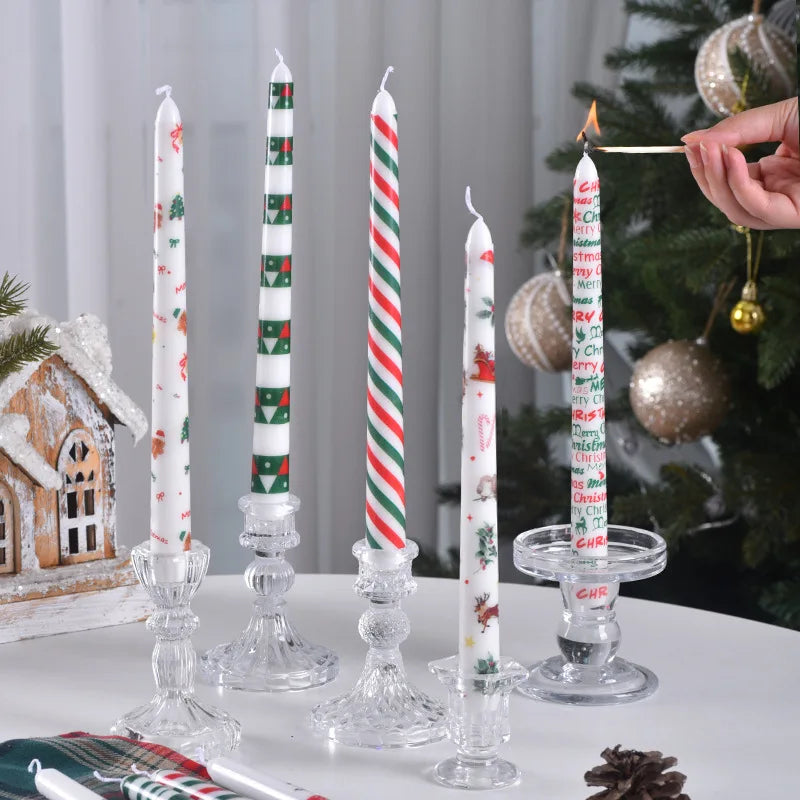 Decorative candles with Christmas designs on a table with a Christmas tree in the background.