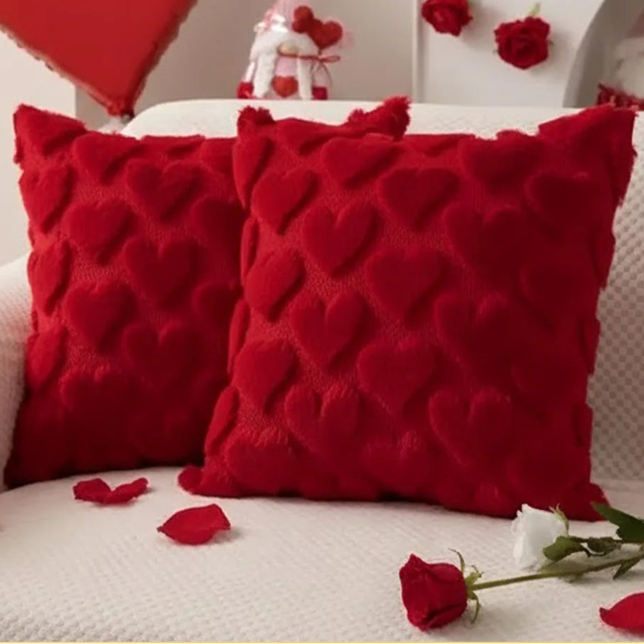 Two red textured heart-patterned pillows on a couch with rose petals and a white rose.