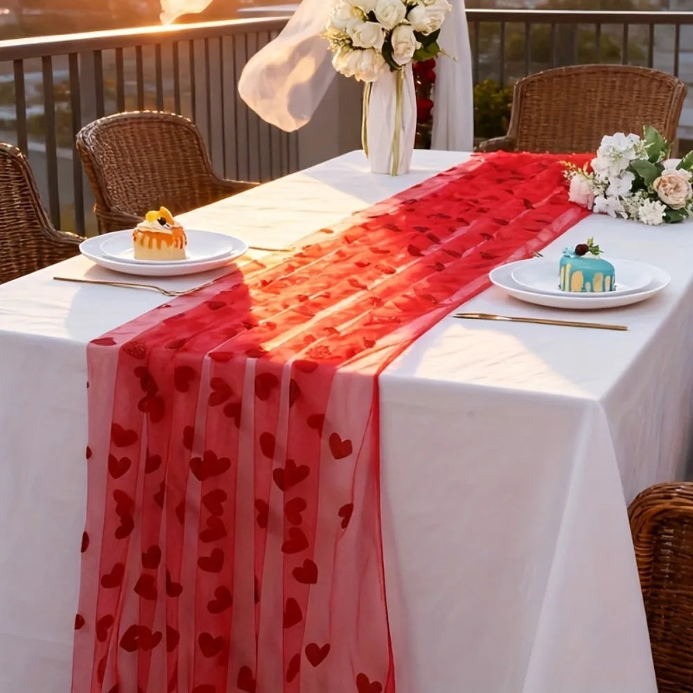 Dining table set with a red heart-patterned runner, white tablecloth, and small cakes on a balcony.