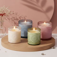Four lit scented candles in textured glass jars in cream, blue, green, and pink, arranged on a round wooden tray against a dusty pink backdrop with a large circular prop and leaf shadows.